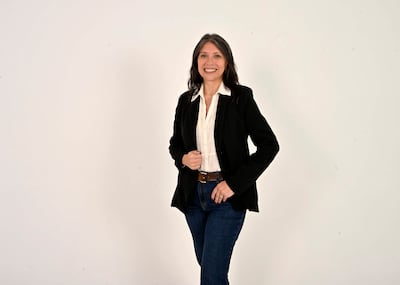 Paola Alarcon sonriendo en blazer negro y blusa blanca, con jeans oscuros, en un fondo blanco, mostrando confianza profesional.