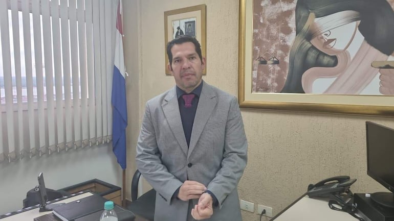 Hombre con traje gris y corbata burdeos interactúa con la cámara en su oficina, con bandera de Paraguay y arte en el fondo.
