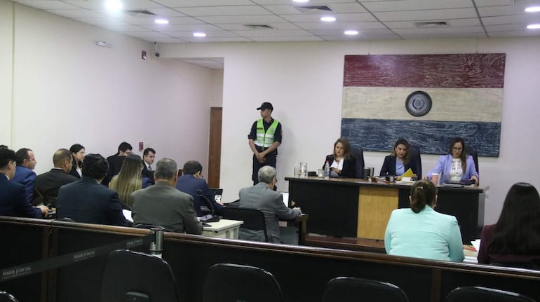 Cinco personas en la mesa central con trajes formales prestando atención, mientras un guardia de seguridad observa al fondo.
