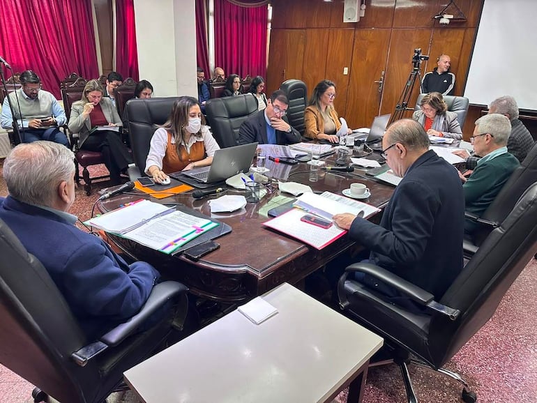 Cecilia Rodríguez presenta en reunión, acompañada por Juan Carlos Frutos y José Emilio Argaña Contreras, en sala de conferencias con decoraciones rojas.
