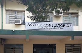 Fachada del Hospital Regional de Pilar con letrero 'ACCESO CONSULTORIOS', árboles y un banco donde se observa una persona con dispositivo móvil.
