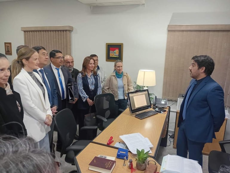 Hombre en traje oscuro hablando frente a un grupo atento en un ambiente profesional, con documentos y una planta en la mesa.
