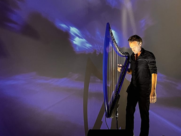 Raoul Moretti con camisa negra, tocando el arpa rodeado de proyecciones en tonos azules y púrpuras.