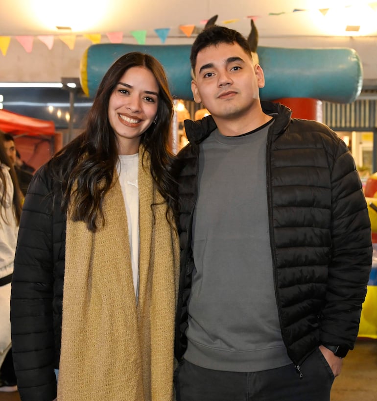 Mujer con abrigo negro y bufanda beige sonriendo junto a hombre con camiseta gris y chaqueta negra en un entorno festivo.