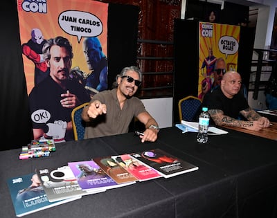 Juan Carlos Tinoco sonríe y señala a la cámara, mientras Octavio Rojas lo observa con una expresión seria en fondo decorado con cómics.
