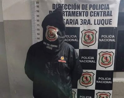 Hombre con chaqueta negra y capucha en comisaría frente a escudo de Policía Nacional, imagen sobria y formal.