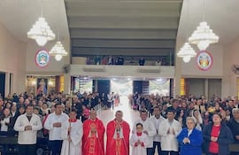 Dos sacerdotes en vestimentas rojas y niños con túnicas blancas sosteniendo velas, rodeados de fieles en actitud reverente.