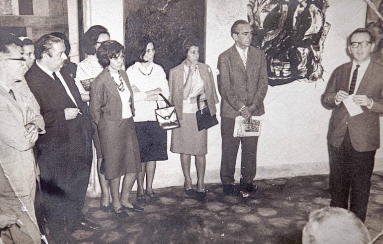 Un hombre sostiene un papel y habla mientras cuatro mujeres lo observan atentamente en un ambiente con arte en las paredes.