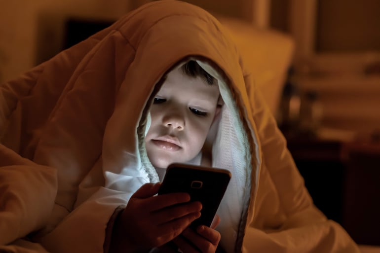 Niño cubierto con manta blanca, concentrado en el teléfono móvil que ilumina su rostro en un ambiente interior.