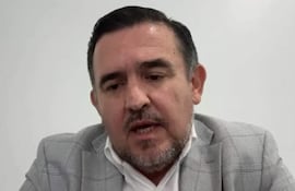 Hombre con barba y cabello corto, vestido con traje gris y camisa blanca, habla sobre un fondo liso y claro.