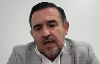 Hombre con barba y cabello corto, vestido con traje gris y camisa blanca, habla sobre un fondo liso y claro.