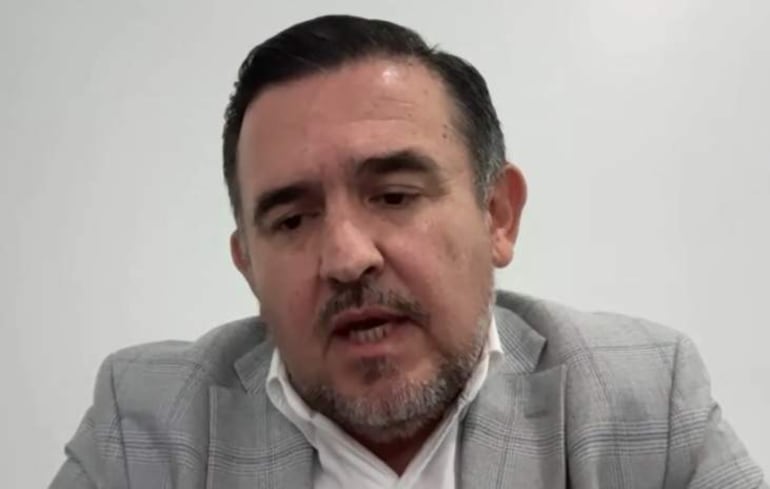 Hombre con barba y cabello corto, vestido con traje gris y camisa blanca, habla sobre un fondo liso y claro.