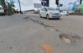 Mujer con camiseta roja usa el teléfono en ruta dañada, mientras otros transeúntes caminan y autos circulan.