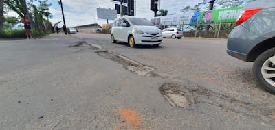 Mujer con camiseta roja usa el teléfono en ruta dañada, mientras otros transeúntes caminan y autos circulan.