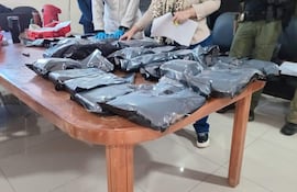 Dos hombres, uno con guantes azules y camisa blanca, sostienen documentos sobre paquetes envueltos en plástico negro en ambiente formal.
