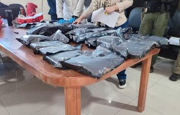 Dos hombres, uno con guantes azules y camisa blanca, sostienen documentos sobre paquetes envueltos en plástico negro en ambiente formal.