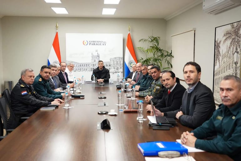 Trece personas en traje formal en reunión, con banderas de Paraguay visibles. Hombre de pie expone frente a otros atentos.