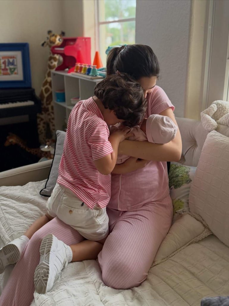 Mujer en pijama rosado con un bebé en brazos, mientras dos niños interactúan en un entorno luminoso y familiar.
