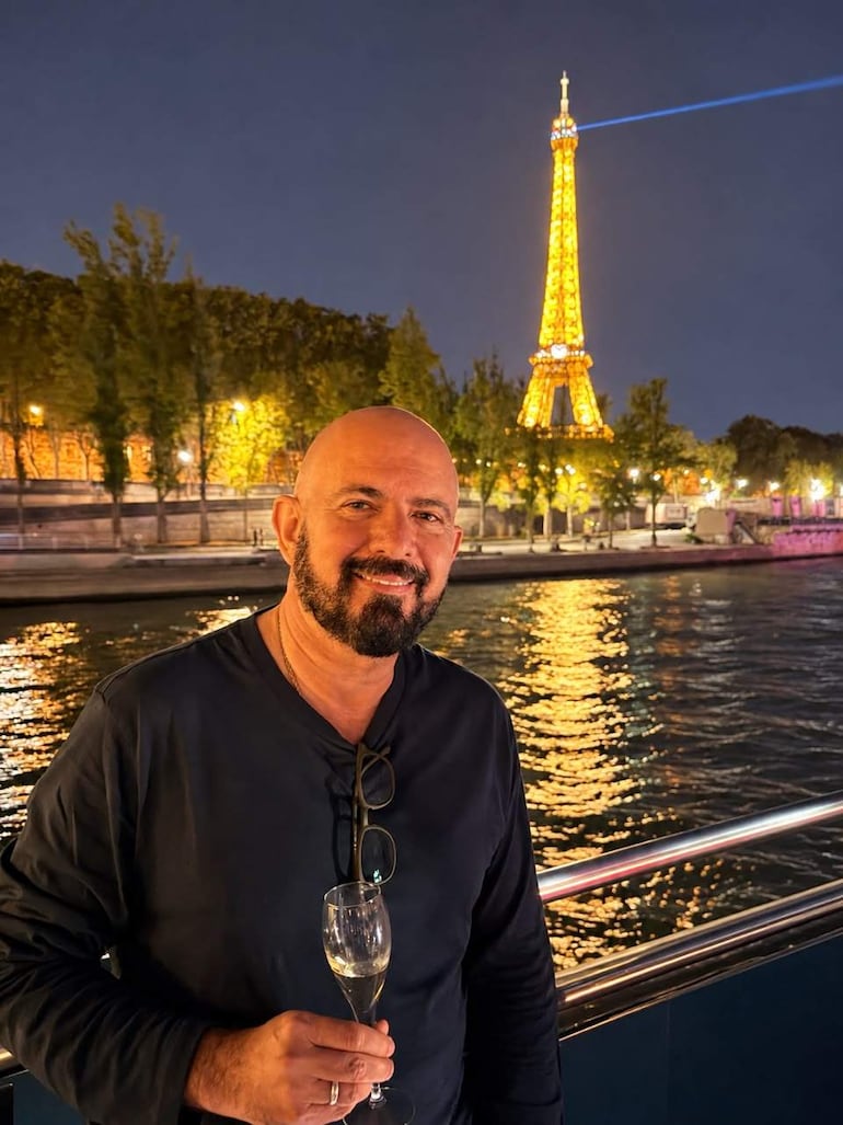 Hombre de pie en camiseta oscura y barba, sonriente, con copa de champagne, frente a la Torre Eiffel iluminada de noche.
