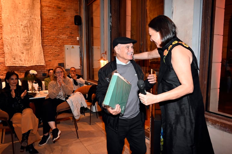 Hombre mayor con gorra sostiene un regalo mientras una mujer de pie en atuendo negro le entrega un reconocimiento en un ambiente festivo.