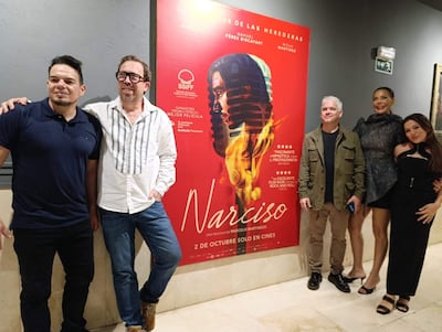 Martinessi en abrigo oliva y Vierci en vestido negro sonríen en el evento de cine con un cartel de 'Narciso' detrás.