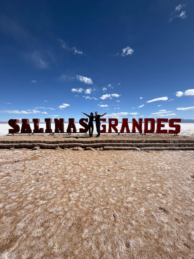 Gloria Vera y Julio Cáceres en Salinas Grandes, sonriendo y con brazos extendidos ante un gran letrero en un paisaje árido.