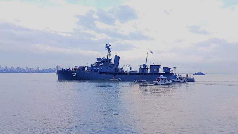 Buque militar C1 en el agua, con minúsculos barcos alrededor y una ciudad al fondo bajo un cielo parcialmente nublado.