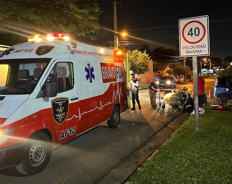 Ambulancia con luces encendidas y dos paramédicos asistiendo a una joven herida en la calle, con un coche azul al fondo.