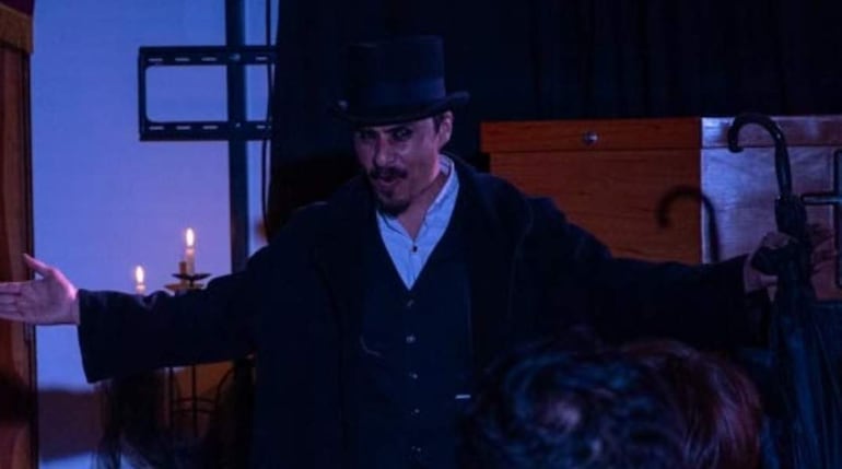 Hombre en vestimenta de época con sombrero de copa y abrigo oscuro, actuando en un ambiente iluminado con velas.