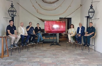 Grupo de seis personas en sillas altas discutiendo sobre fiestas patrias, con hombre de barba y gafas hablando y bandera paraguaya al fondo.