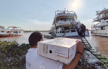 Hombre con camiseta blanca sostiene caja electoral en puerto, mientras varias personas observan desde un barco.