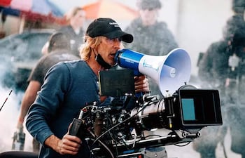 Michael Bay, con cabello largo y camiseta de manga larga, dirige un rodaje al aire libre con un megáfono en mano, rodeado de humo.