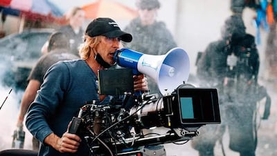 Michael Bay, con cabello largo y camiseta de manga larga, dirige un rodaje al aire libre con un megáfono en mano, rodeado de humo.