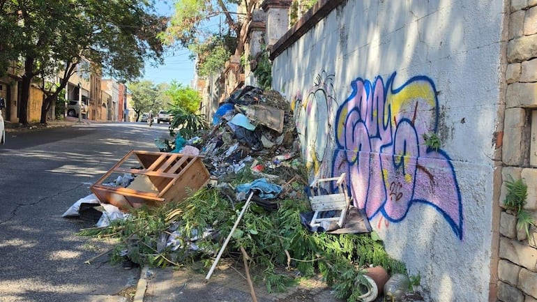Montón de basura, incluyendo plásticos y un mueble, en una calle con grafitis coloridos y vegetación al fondo.