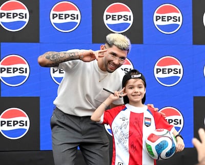 Joven rubio con tatuajes y niño con camiseta de selección paraguaya posan felices, sosteniendo un balón de fútbol decorado.