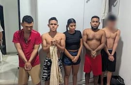 Cinco jóvenes colombianos en habitación sencilla, dos sin camiseta y otros con ropa casual, luciendo serios y alineados.