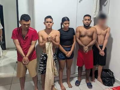 Cinco jóvenes colombianos en habitación sencilla, dos sin camiseta y otros con ropa casual, luciendo serios y alineados.