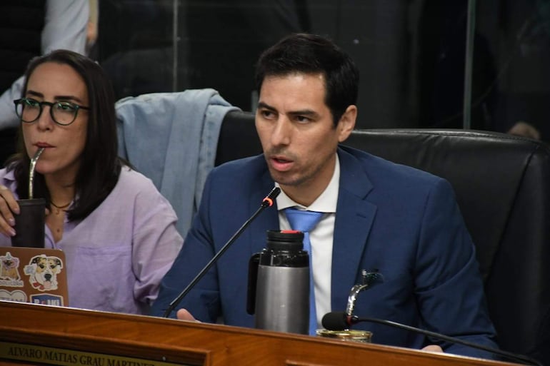 Alvaro Grau en traje azul con corbata blanca habla mientras Jazmin Galeano, en blusa lila y gafas, escucha atentamente.