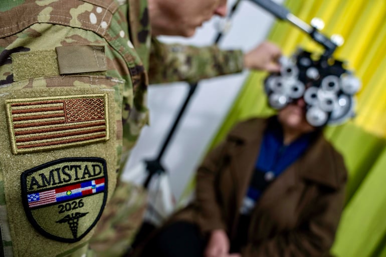 Soldado en uniforme camuflado con parche 'AMISTAD 2026', observa a un paciente con dispositivo óptico en ambiente interior.