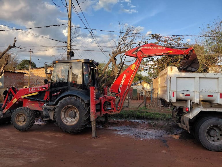 Tractor rojo opera en zona urbana, cargando materiales en camión blanco bajo un cielo nublado.