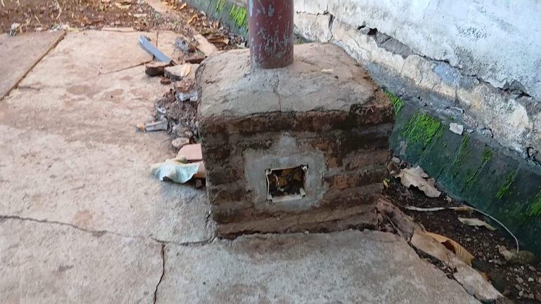 Estructura de cemento deteriorada con un poste metálico en un entorno urbano lleno de escombros y grietas en el pavimento.