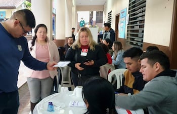 Mujer con blusa rosa y chaqueta clara recibe un documento de un hombre robusto con gafas y camiseta azul, rodeados de otros participantes.