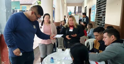 Mujer con blusa rosa y chaqueta clara recibe un documento de un hombre robusto con gafas y camiseta azul, rodeados de otros participantes.