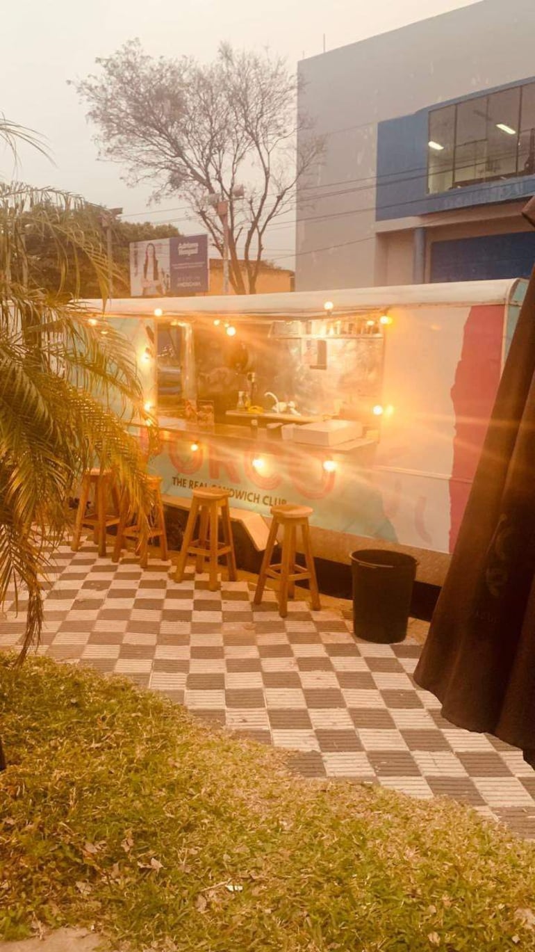 Carro de comida decorado con luces, ventanilla para preparación de alimentos, y sillas de madera vacías en un entorno al aire libre.