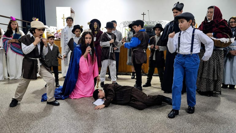 Niña con vestido rosa y capa azul sostiene un micrófono mientras otros niños actúan en un ambiente festivo escolar.