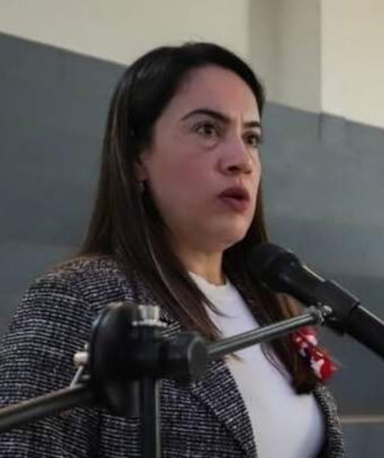 Mujer de blusa blanca y saco de cuadros hablando frente a un micrófono en un ambiente sobrio.