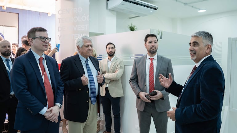 Hombres con trajes y corbatas discuten en un ambiente interior, con un hombre mayor hablando y otros atentos en la conversación.