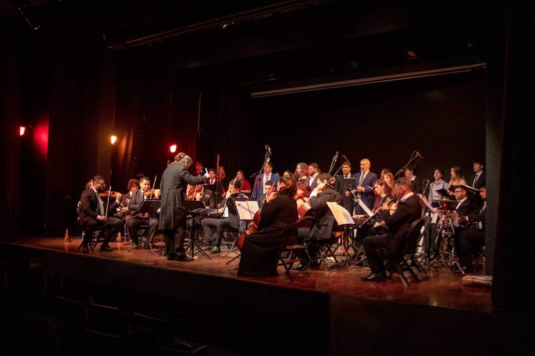 Grupo de músicos en trajes oscuros, director al frente, tocando en un ambiente teatral iluminado con luces rojas.