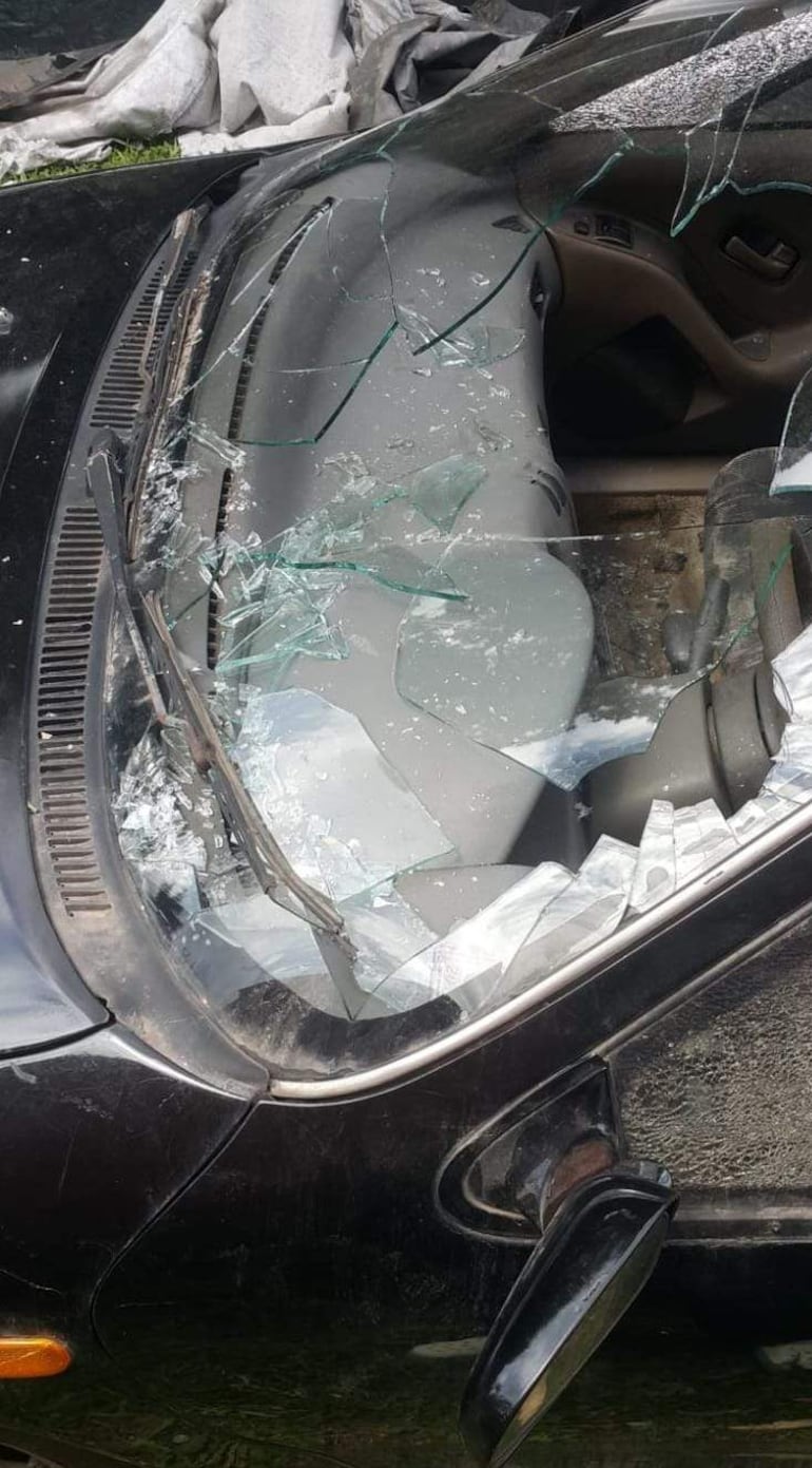 Automóvil negro dañado, con parabrisas roto y vidrios esparcidos, en un entorno con césped y una tela de fondo.