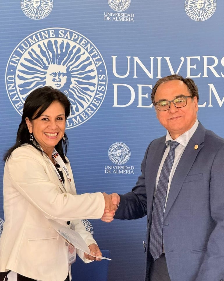 Mujer con blazer blanco y hombre en traje oscuro se estrechan la mano, ambos sonrientes en un entorno formal con fondo azul.